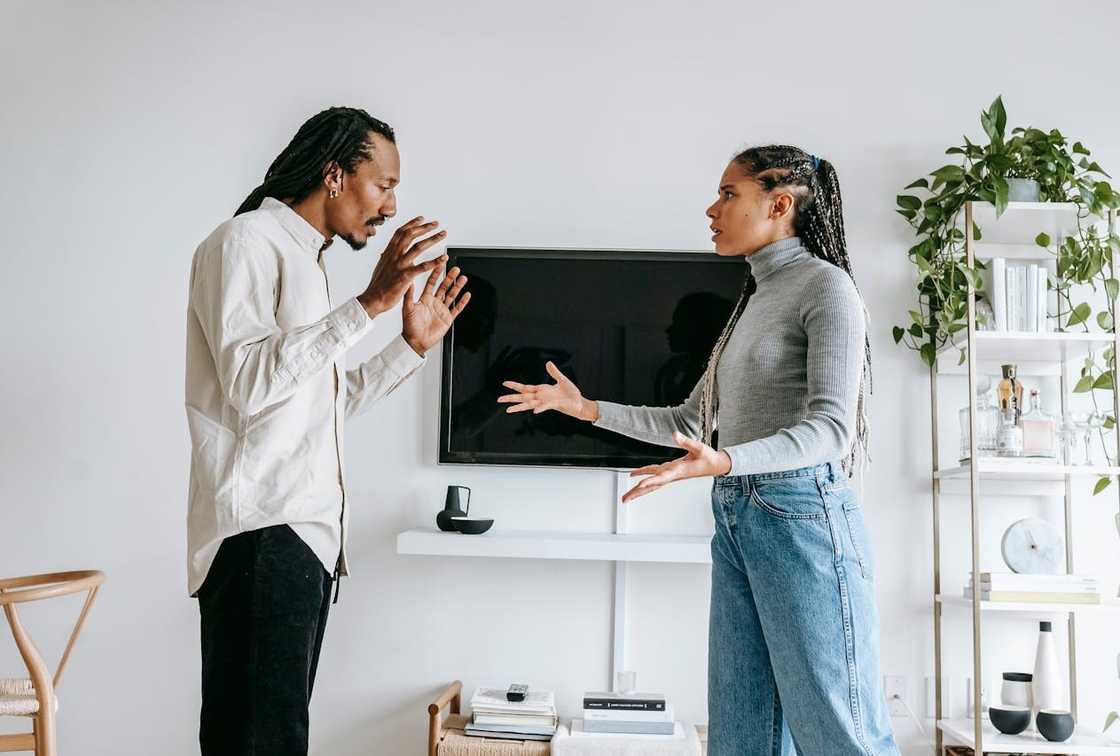 A man and woman stand in a living room arguing with raised hands and tense expressions.