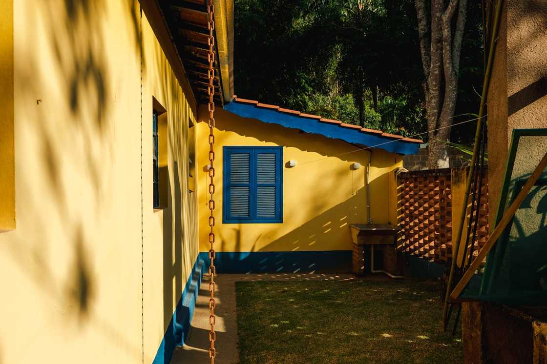 Small yellow house with blue window shutters and a shaded garden area.