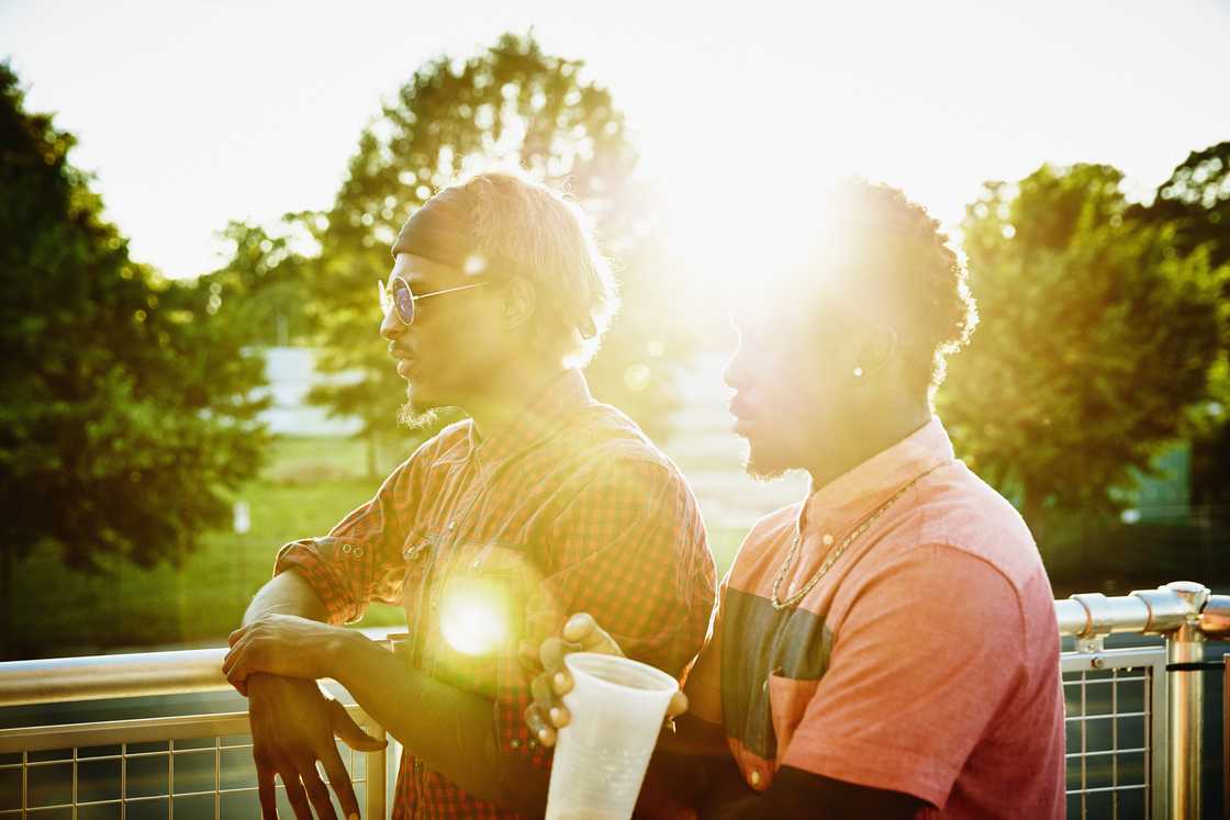 Two people stand on a railing at sunset, partially obscured by bright sunlight.