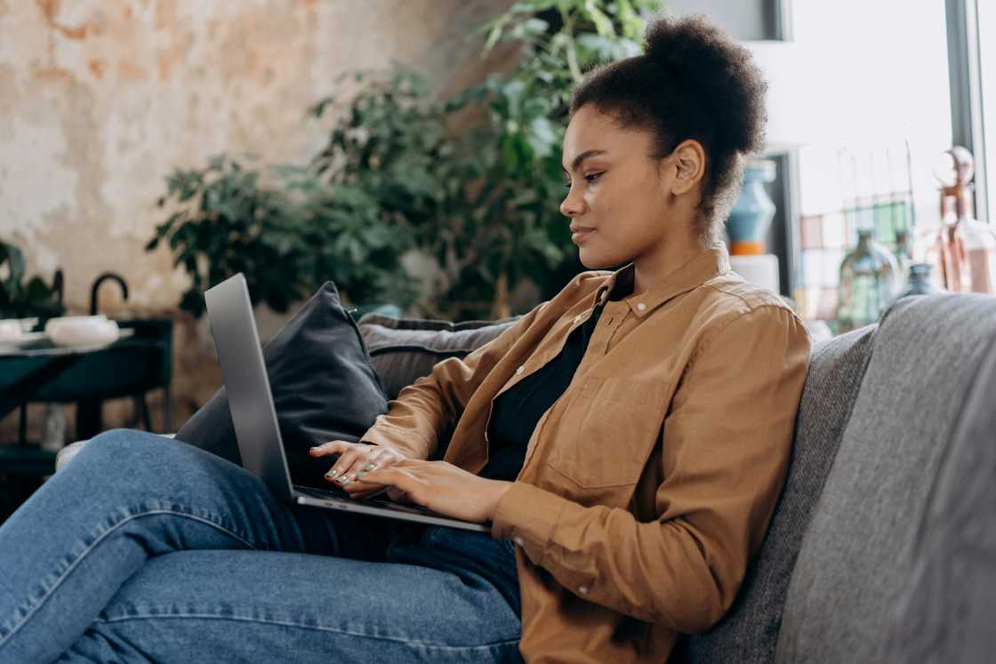 A woman in denim pants and a brown jacket is using a laptop on a grey sofa A woman in denim pants and a brown jacket is using a laptop on a grey sofa