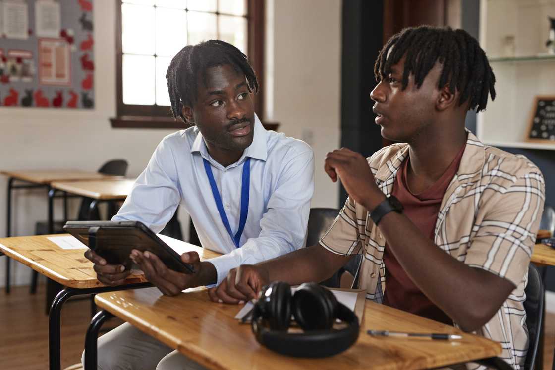 A man holding a tablet sits beside a student wearing a plaid shirt, both talking, with headphones resting on the desk between them.