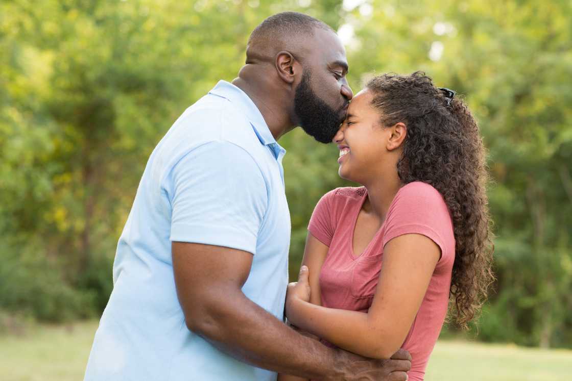 Man kisses a smiling young girl on the forehead in a green outdoor setting. Man kisses a smiling young girl on the forehead in a green outdoor setting.
