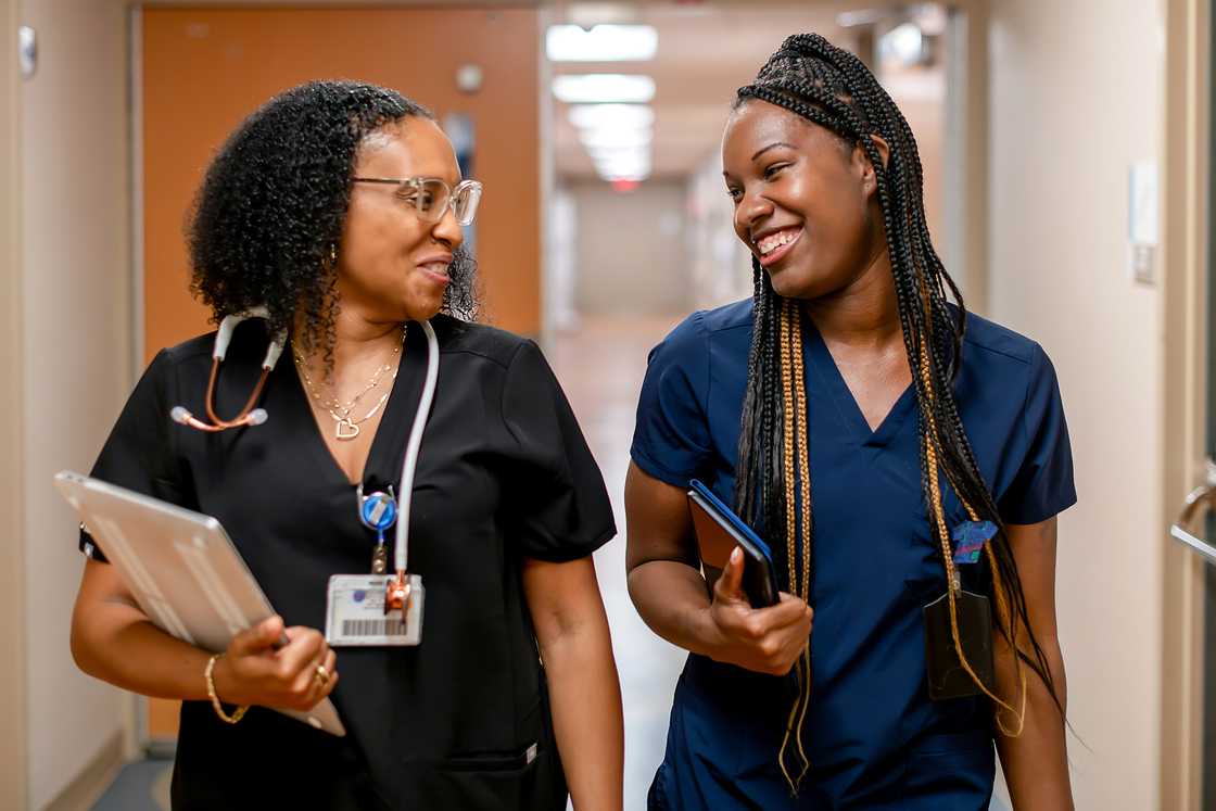 A woman in black scrubs with a stethoscope and an ID badge walks beside a woman in navy scrubs with long braided hair, both smiling at each other. A woman in black scrubs with a stethoscope and an ID badge walks beside a woman in navy scrubs with long braided hair, both smiling at each other.