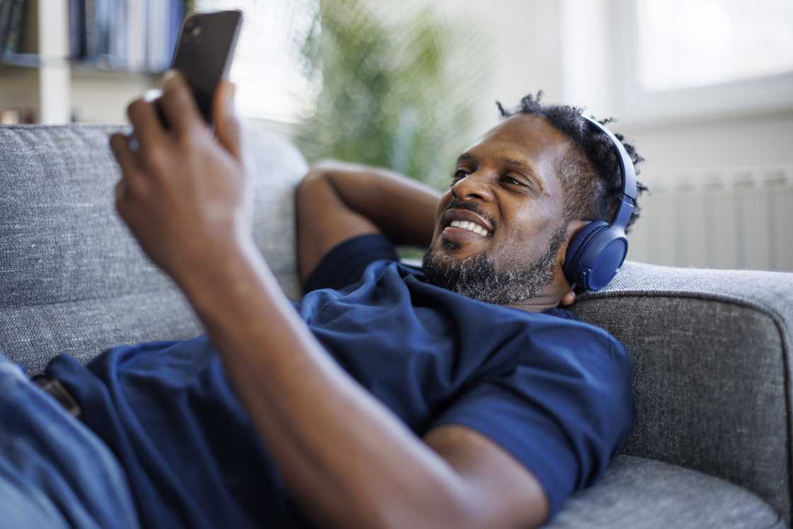 A man with Bluetooth headphones using a mobile phone and relaxing on a sofa.