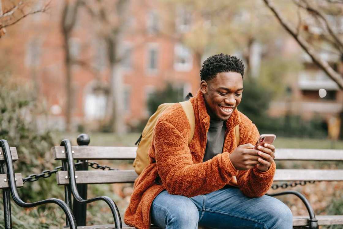 A happy young man wearing denim pants and a brown jacket is using a smartphone