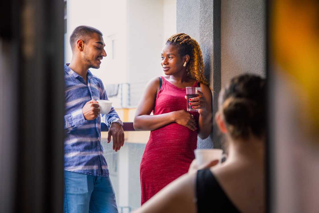 Three people on a balcony, two talking, one seated with a cup.