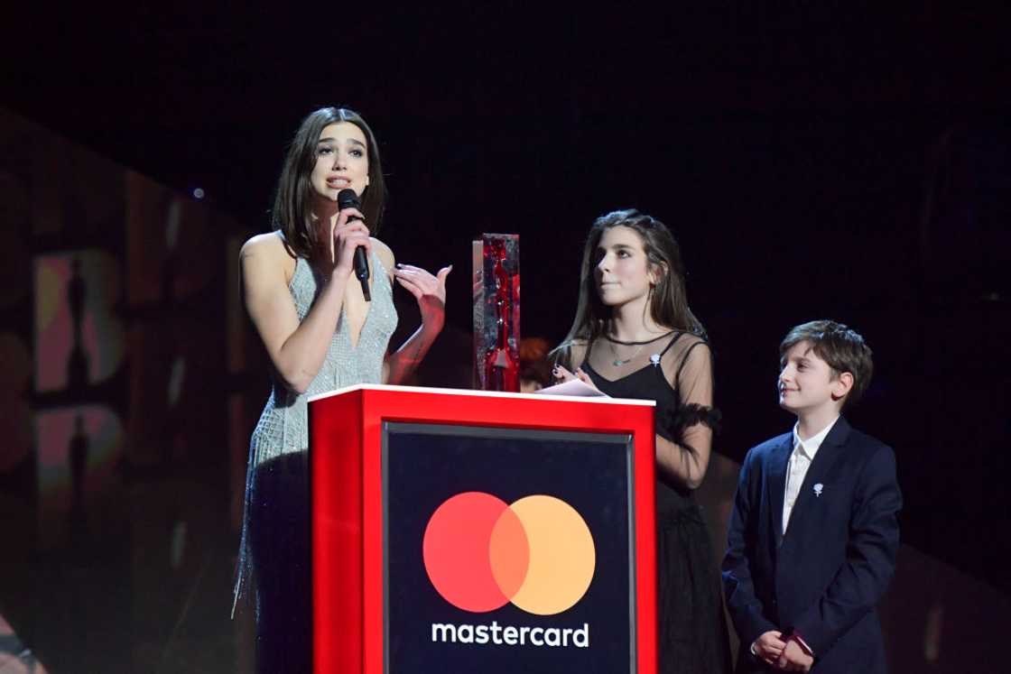 A woman in a sequined dress speaks into a microphone at a red Mastercard podium beside a girl and boy. A woman in a sequined dress speaks into a microphone at a red Mastercard podium beside a girl and boy.