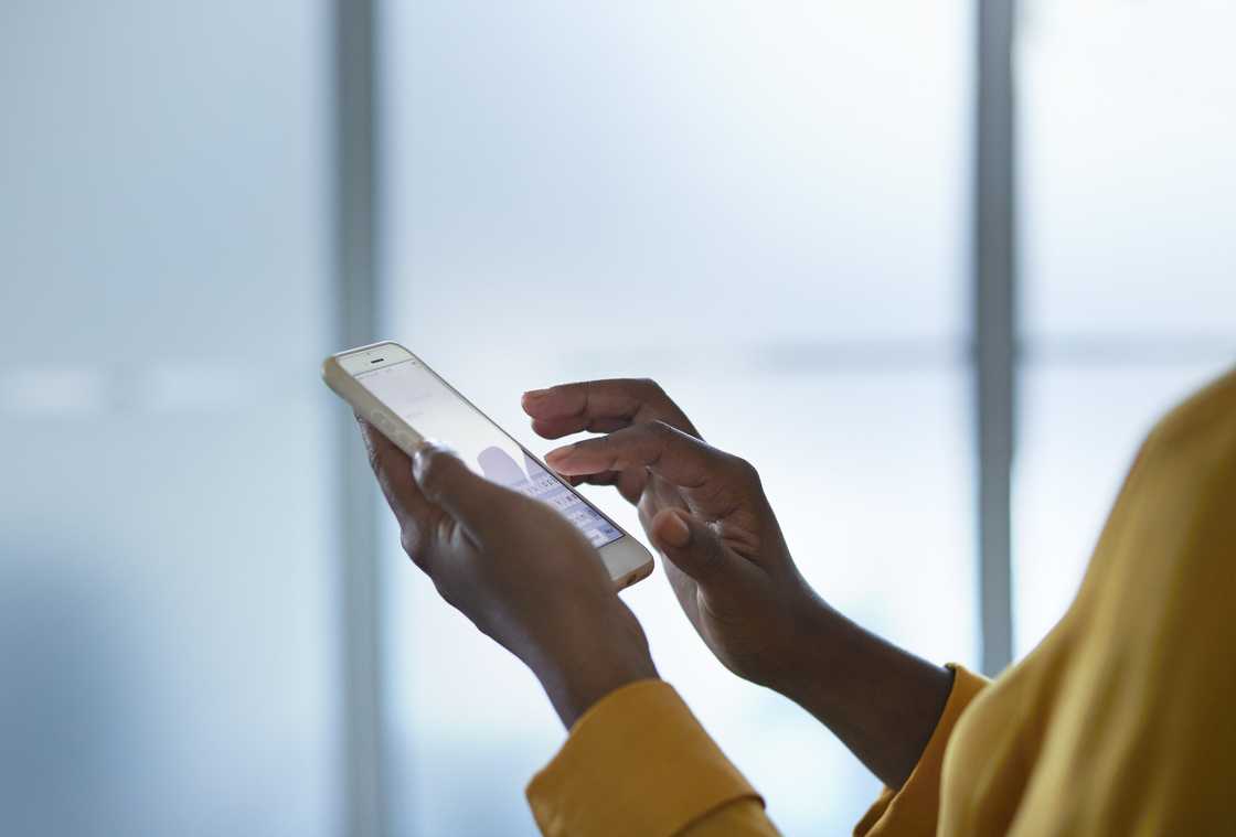 A close-up of hands in a mustard-yellow top typing on a white smartphone against a bright, blurred background.
