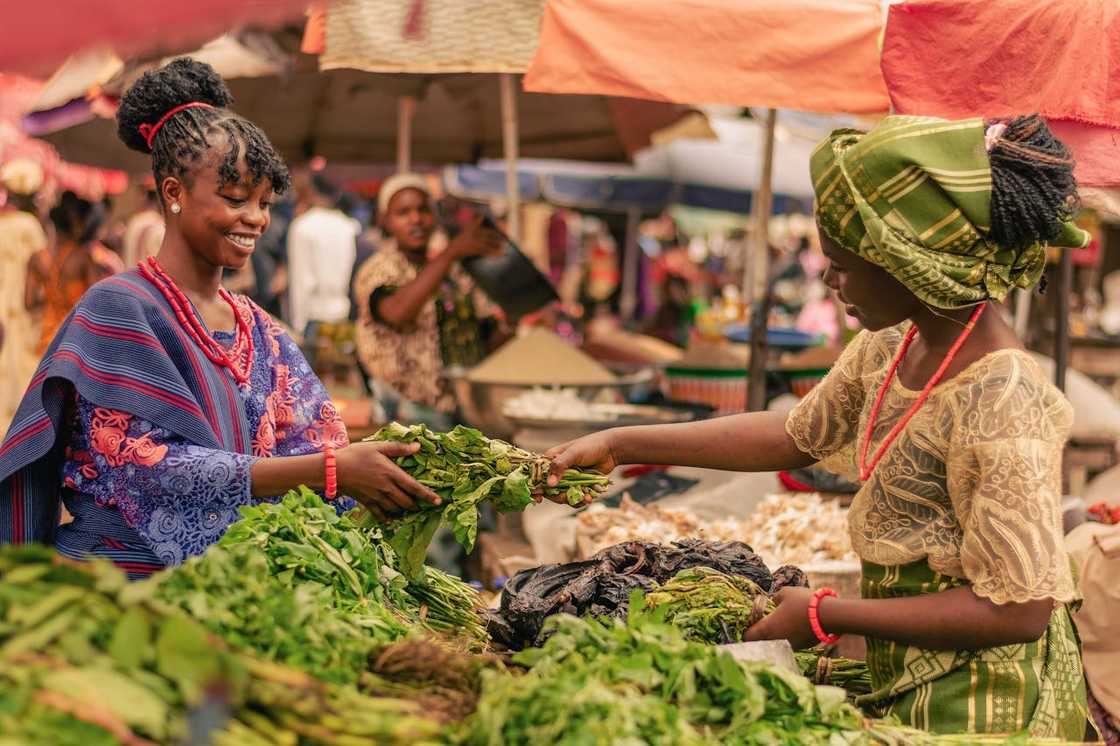 Two women exchange leafy vegetables at an outdoor market. Two women exchange leafy vegetables at an outdoor market.