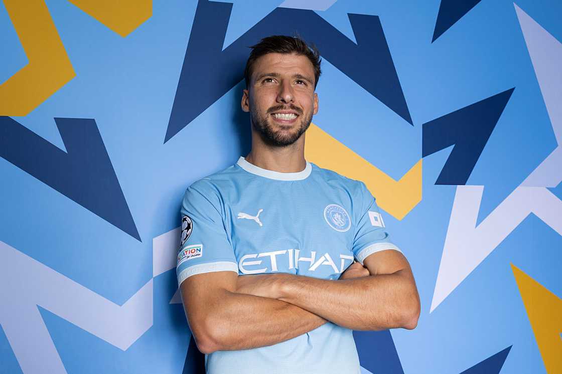 Rúben Dias, in the sky-blue Manchester City home kit, poses with arms folded against a blue patterned backdrop. Rúben Dias, in the sky-blue Manchester City home kit, poses with arms folded against a blue patterned backdrop.