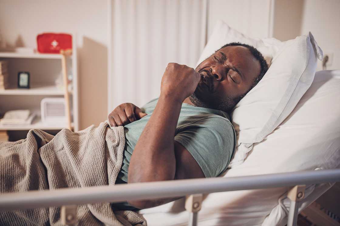 A person lying in a hospital bed with a beige blanket, appearing unwell