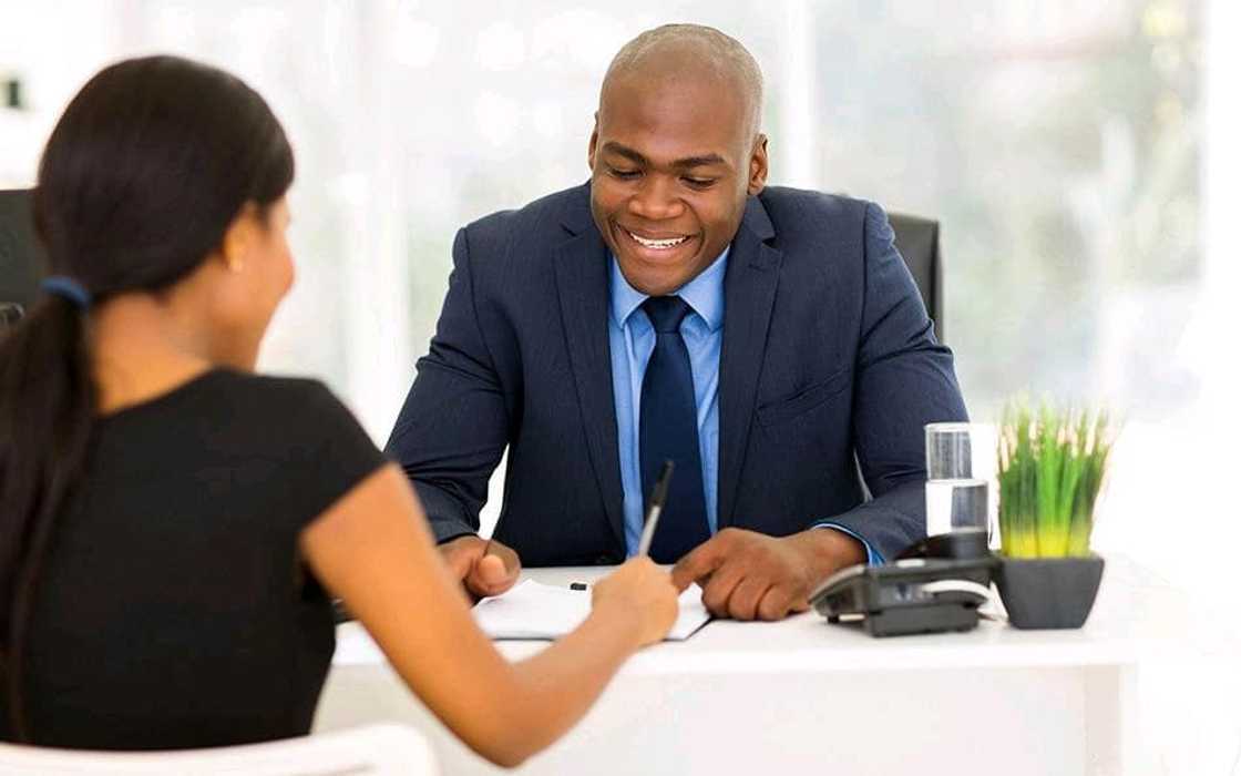 A man in a suit guides a woman as she writes on a document an an office setting.