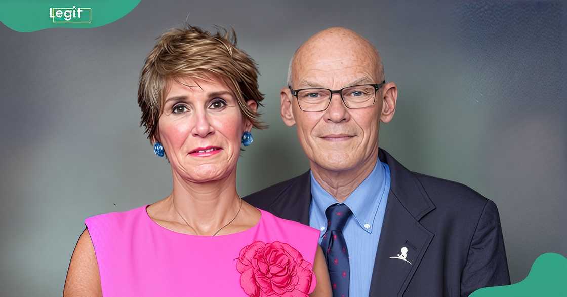James Carville's wife poses in front of him in a pink dress against a grey background