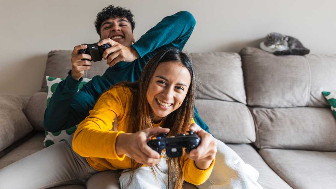 A man in a green hoodie and a woman in a yellow top passionately play video games on a grey couch.