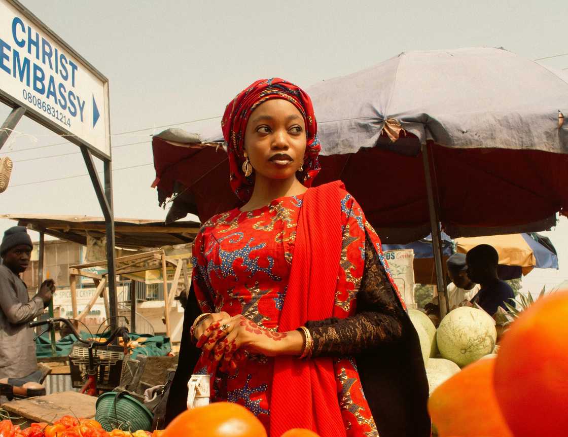 A woman in a red dress and headscarf stands in a busy outdoor market.