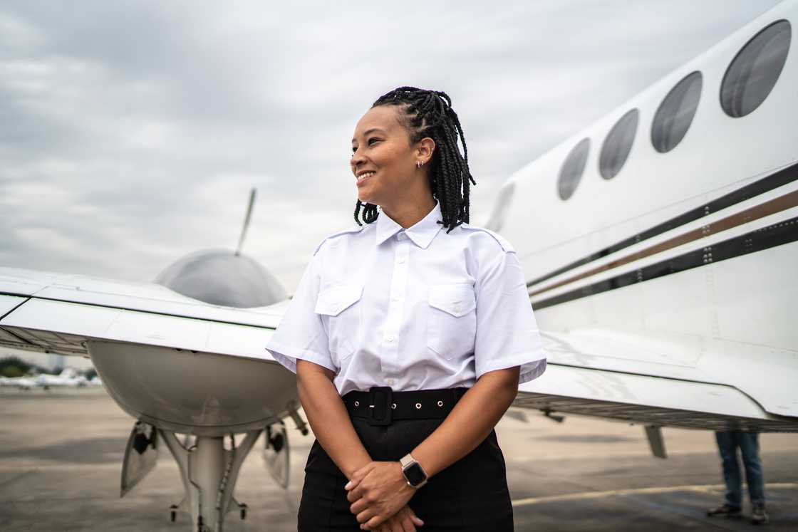 A woman in a white pilot's shirt and black pants stands in front of a small jet, smiling as she looks off to the side. A woman in a white pilot's shirt and black pants stands in front of a small jet, smiling as she looks off to the side.