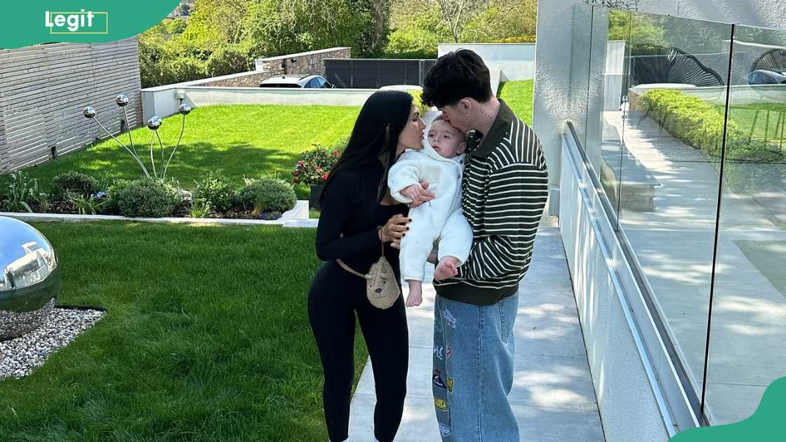 Cam and Emily standing outdoors next to a modern building with their child Cam and Emily standing outdoors next to a modern building with their child