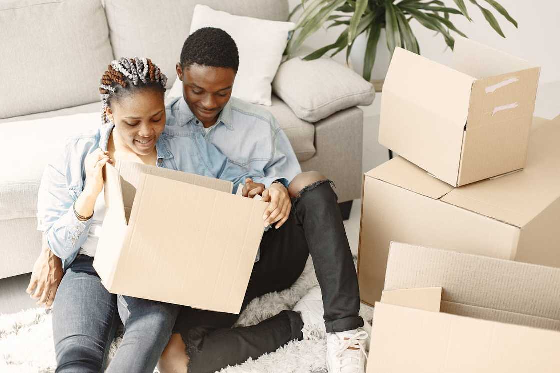 A couple sits on the floor opening a cardboard box while unpacking in a living room.