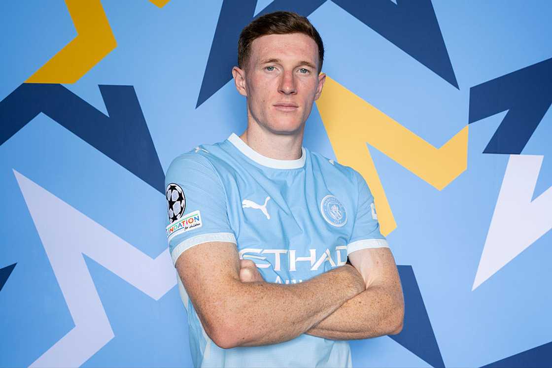 Elliot Anderson, in the sky-blue Manchester City home kit, poses against a blue patterned backdrop. Elliot Anderson, in the sky-blue Manchester City home kit, poses against a blue patterned backdrop.