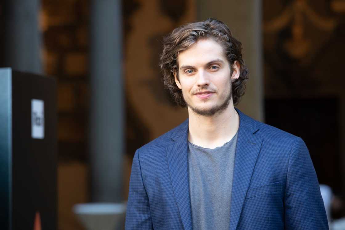 A man with curly brown hair and light eyes wears a blue blazer over a gray T-shirt.