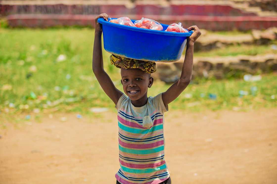 A young boy carrying a basin of goods on his head outdoors.
