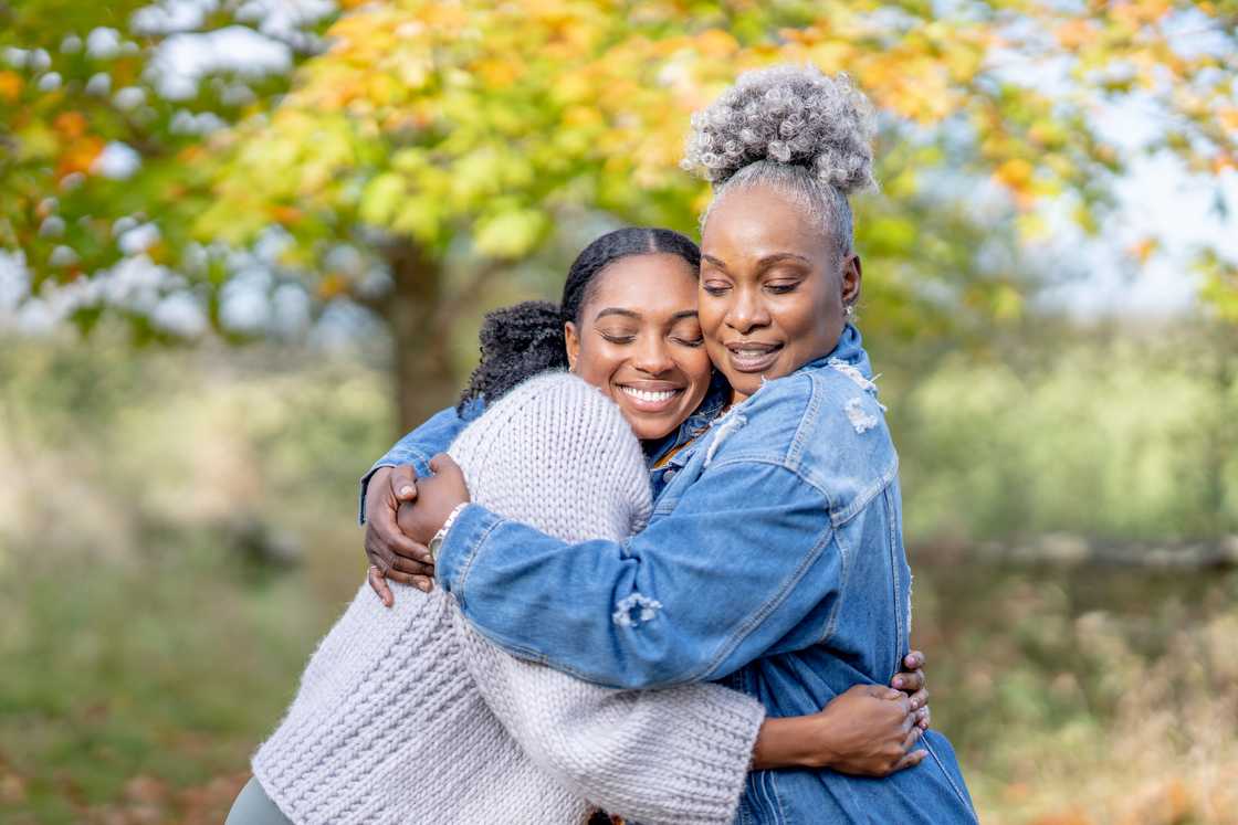 Two people embrace outdoors, one in a grey sweater and the other in a denim jacket.