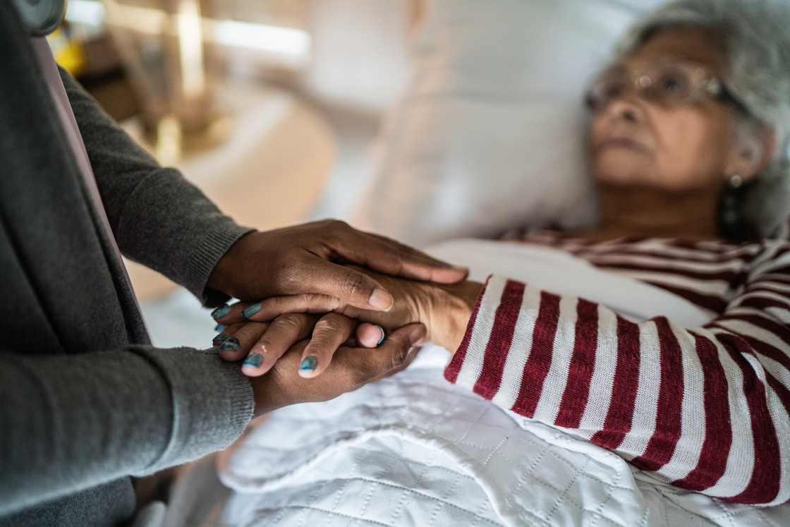Older person lies in a hospital bed while another individual gently holds their hand in a comforting gesture. Older person lies in a hospital bed while another individual gently holds their hand in a comforting gesture.
