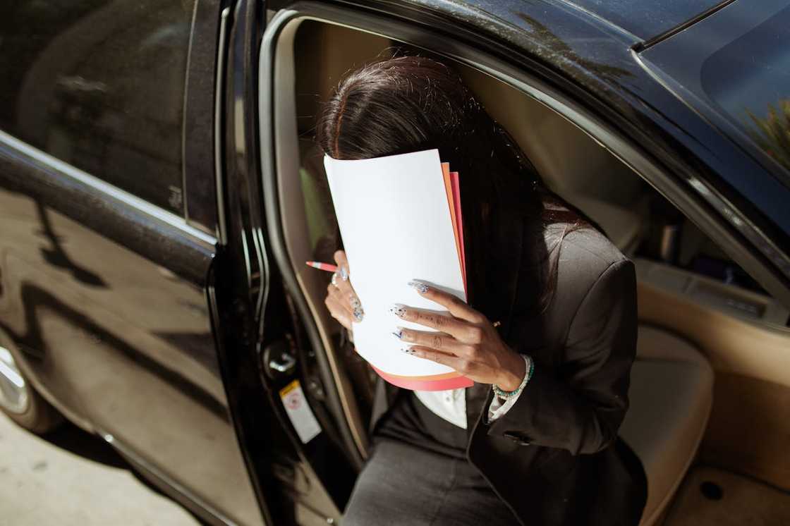 A person stepping out of a car while covering their face with documents.