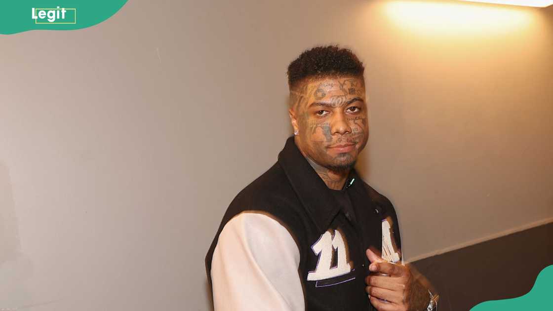 A man with extensive black face tattoos and short curly hair poses at an indoor event