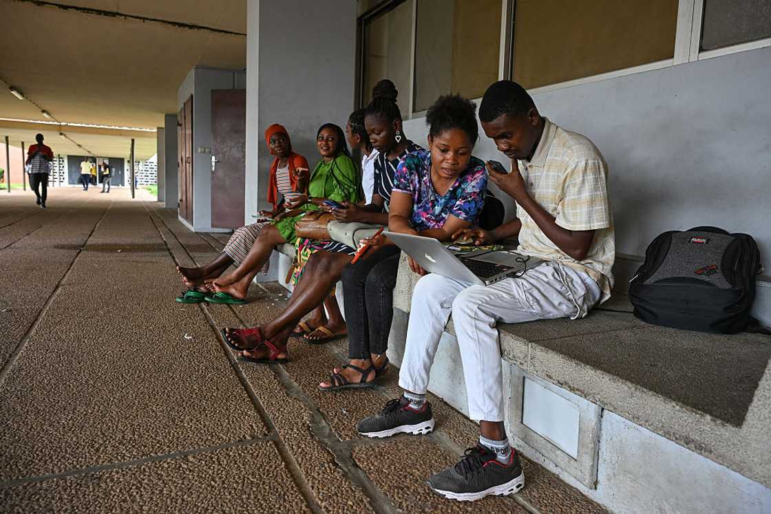 A young man holds a laptop while two young women look at the screen together outside a glass-fronted building. A young man holds a laptop while two young women look at the screen together outside a glass-fronted building.