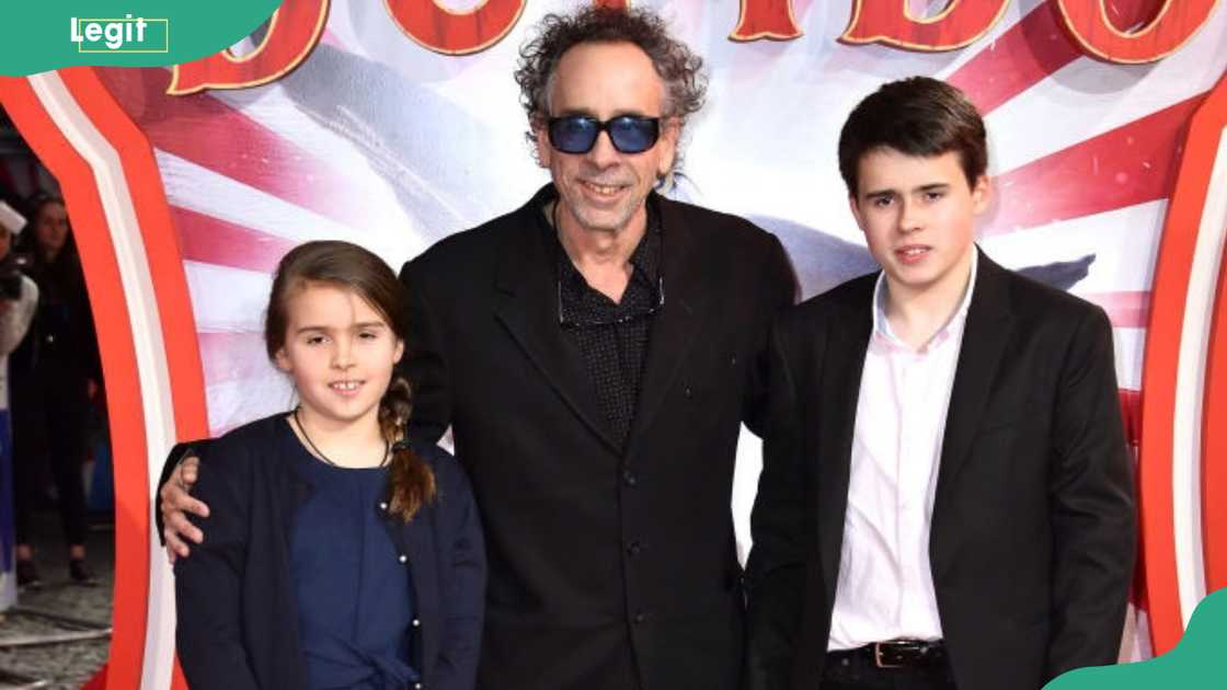 A curly-haired man in sunglasses stands between a young girl and a teenage boy in front of a Dumbo-themed backdrop.