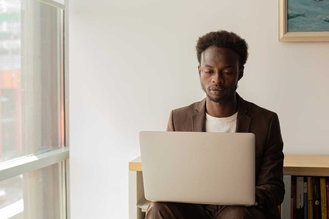 A man sits by a window working on a laptop.