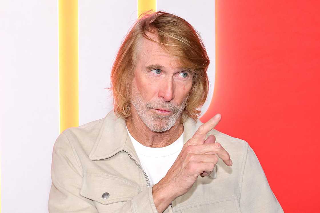 Michael Bay wearing a beige jacket over a white t-shirt, pointing with his hand and looking to the side against a colorful backdrop. Michael Bay wearing a beige jacket over a white t-shirt, pointing with his hand and looking to the side against a colorful backdrop.