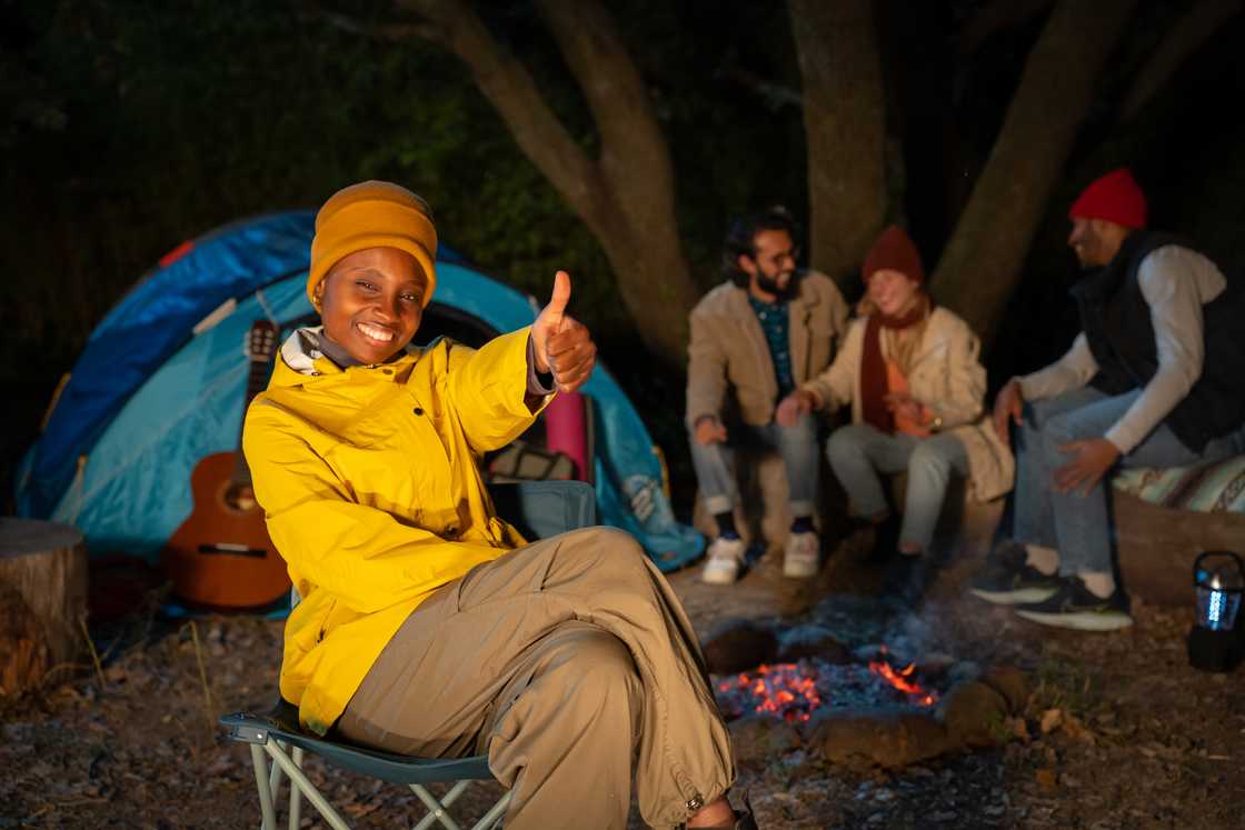 a young black girl giving thumbs up happy with her friends in the background camping outdoors at night.