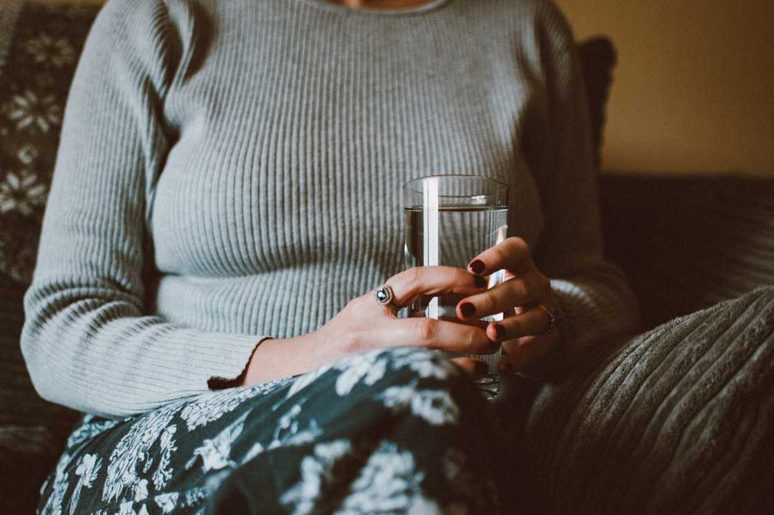 A woman sits holding a glass of water, looking thoughtful.