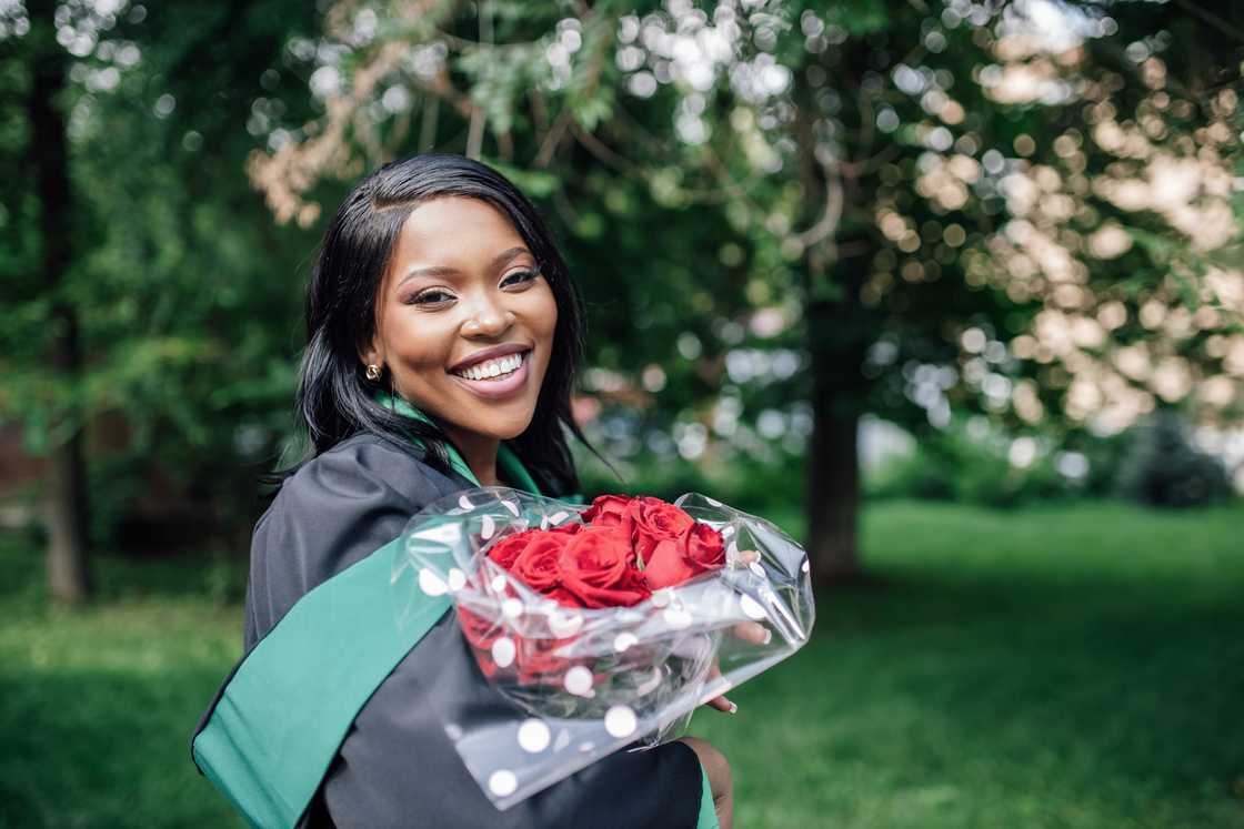 A lady in graduation gown standing in public park holding bouquet of red roses A lady in graduation gown standing in public park holding bouquet of red roses