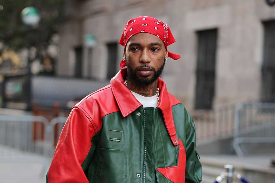 A man in a red bandana and red-and-green leather jacket stands outdoors on a city street. A man in a red bandana and red-and-green leather jacket stands outdoors on a city street.