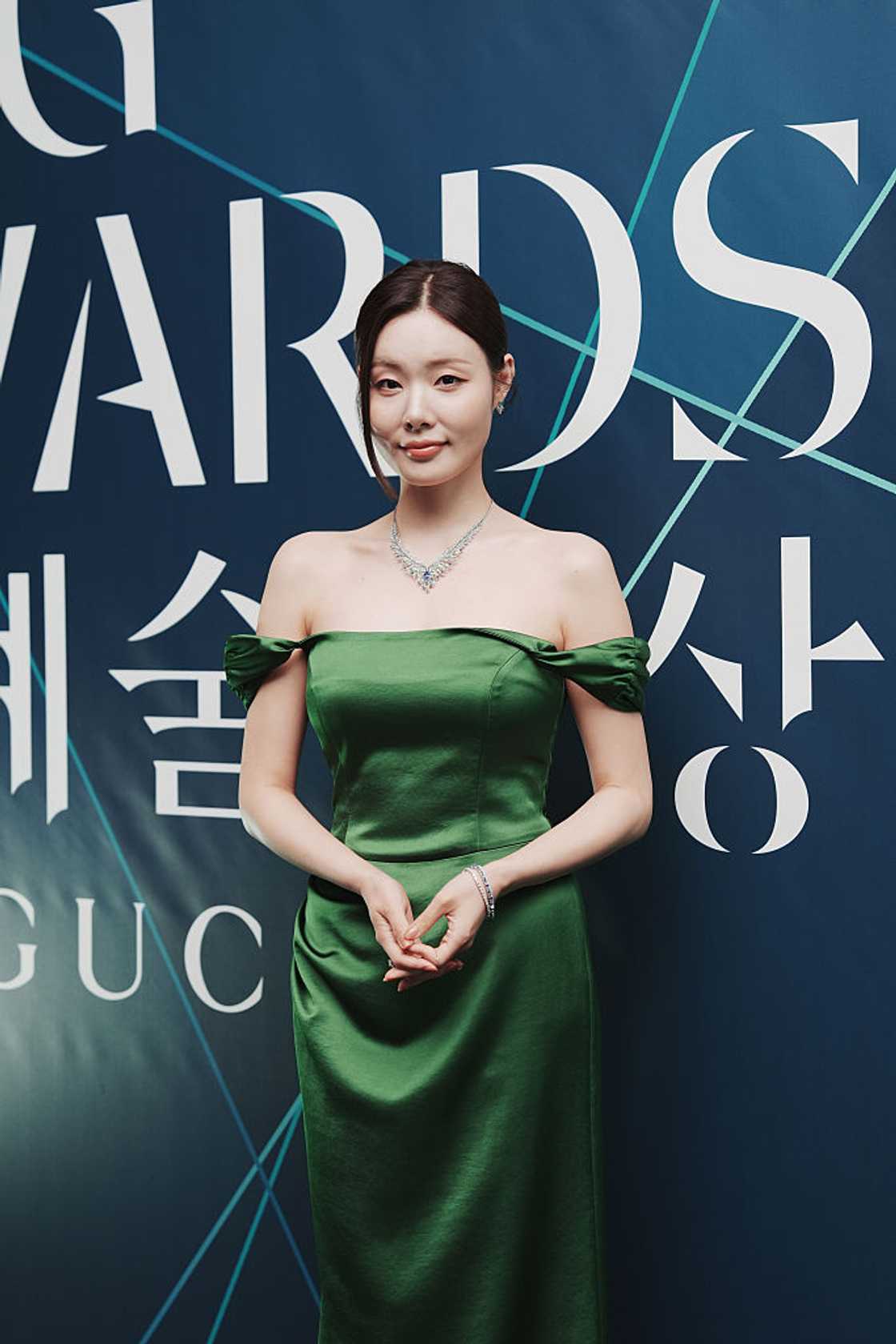 A woman in an off-shoulder green satin gown poses in front of an awards event backdrop. A woman in an off-shoulder green satin gown poses in front of an awards event backdrop.