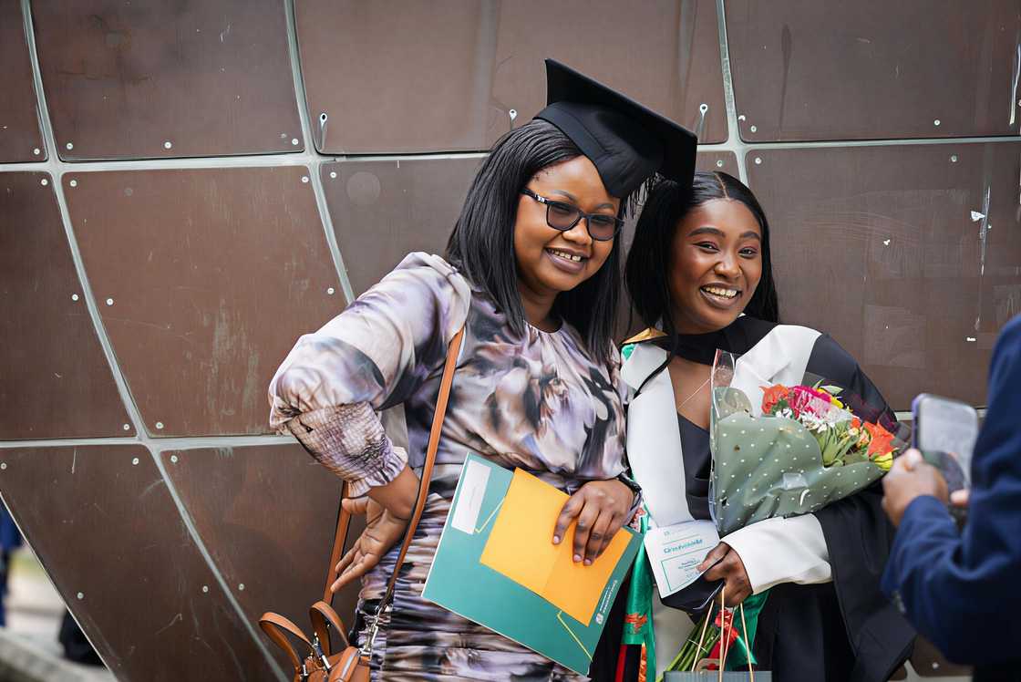 Two smiling ladies, one in a graduation cap and gown holding a bouquet of flowers