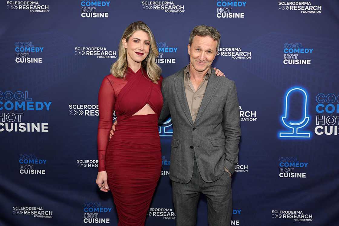 A woman in a cutout red dress stands with a man in a gray suit at a step-and-repeat. A woman in a cutout red dress stands with a man in a gray suit at a step-and-repeat.