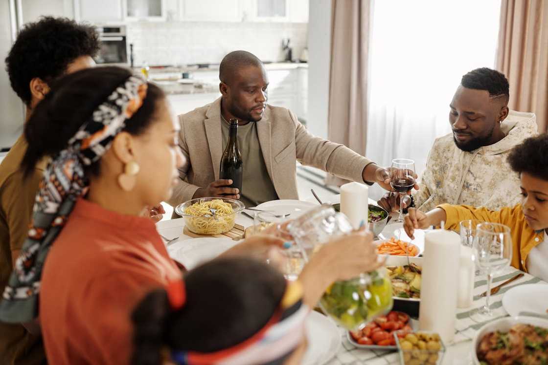 A family eats together at a dining table while a man pours wine for another guest. A family eats together at a dining table while a man pours wine for another guest.
