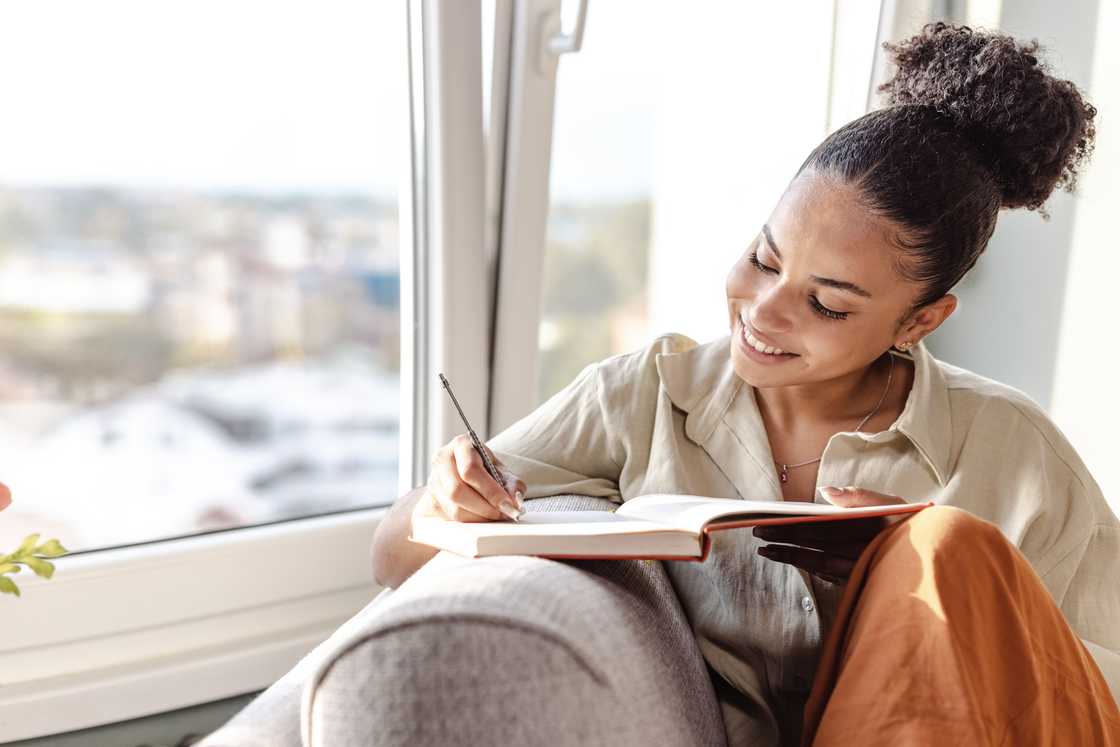 Person sits on a couch near a window, smiling while writing in a notebook.