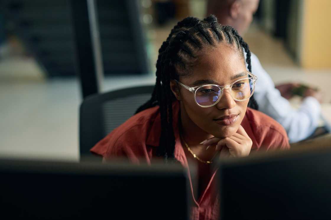 A woman with braided hair and clear-framed glasses rests her chin on her hand while looking intently at a monitor.