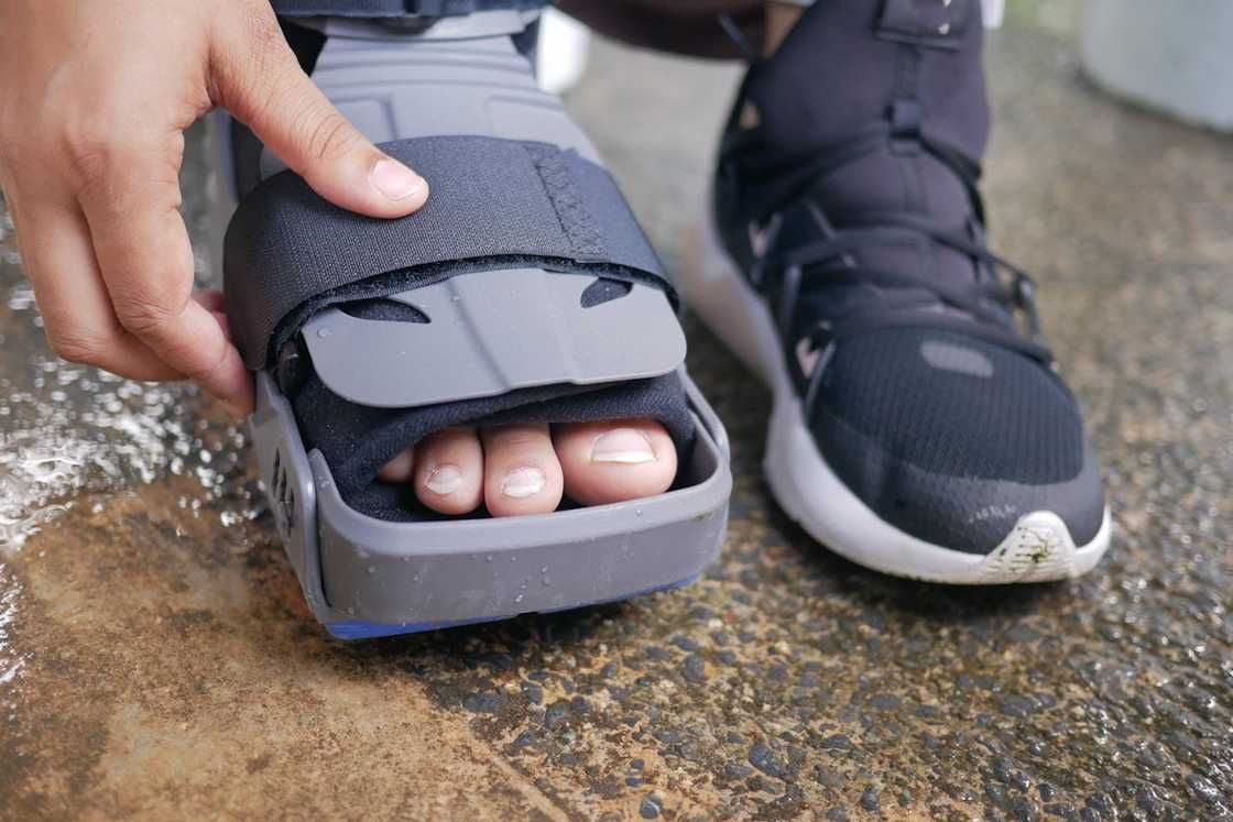 A person adjusts a medical walking boot on one foot while wearing a trainer on the other. A person adjusts a medical walking boot on one foot while wearing a trainer on the other.