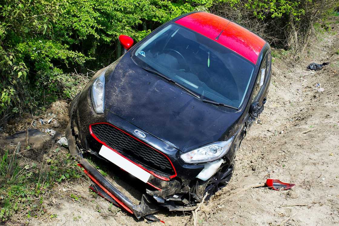 A damaged black car with a red roof resting off the road in a dirt ditch.