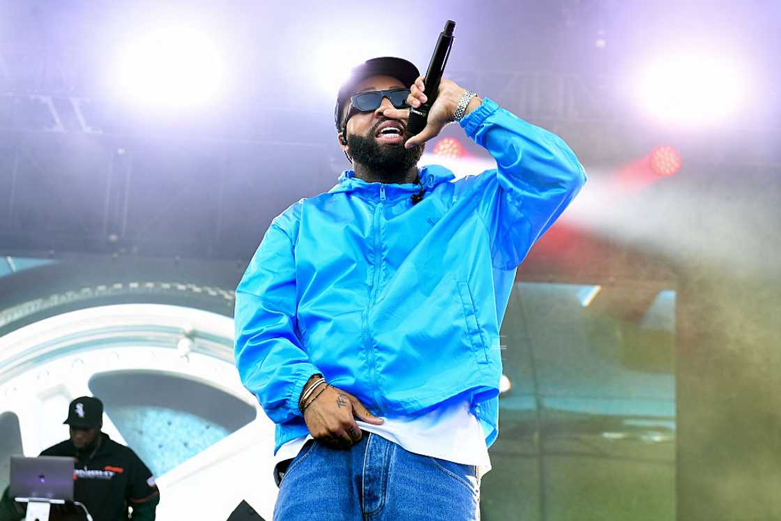 Larry June in a blue windbreaker and sunglasses raises a microphone while performing on stage. Larry June in a blue windbreaker and sunglasses raises a microphone while performing on stage.