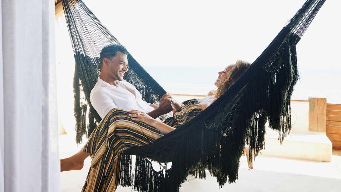 A man in a white shirt and a woman in a multicoloured outfit lounge in a black hammock in a bright, airy space.