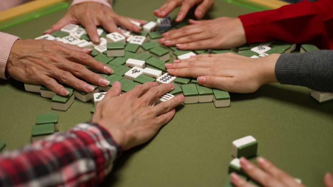 A closeup view of hands mixing tiles on the table with green cloth during a mahjong game.