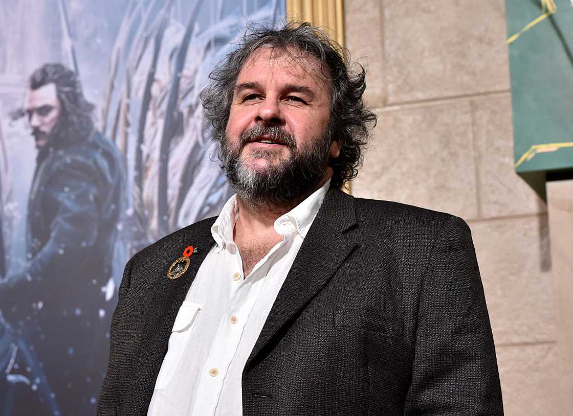 Peter Jackson wearing a dark blazer over a white shirt, smiling in front of a movie poster at a premiere event. Peter Jackson wearing a dark blazer over a white shirt, smiling in front of a movie poster at a premiere event.