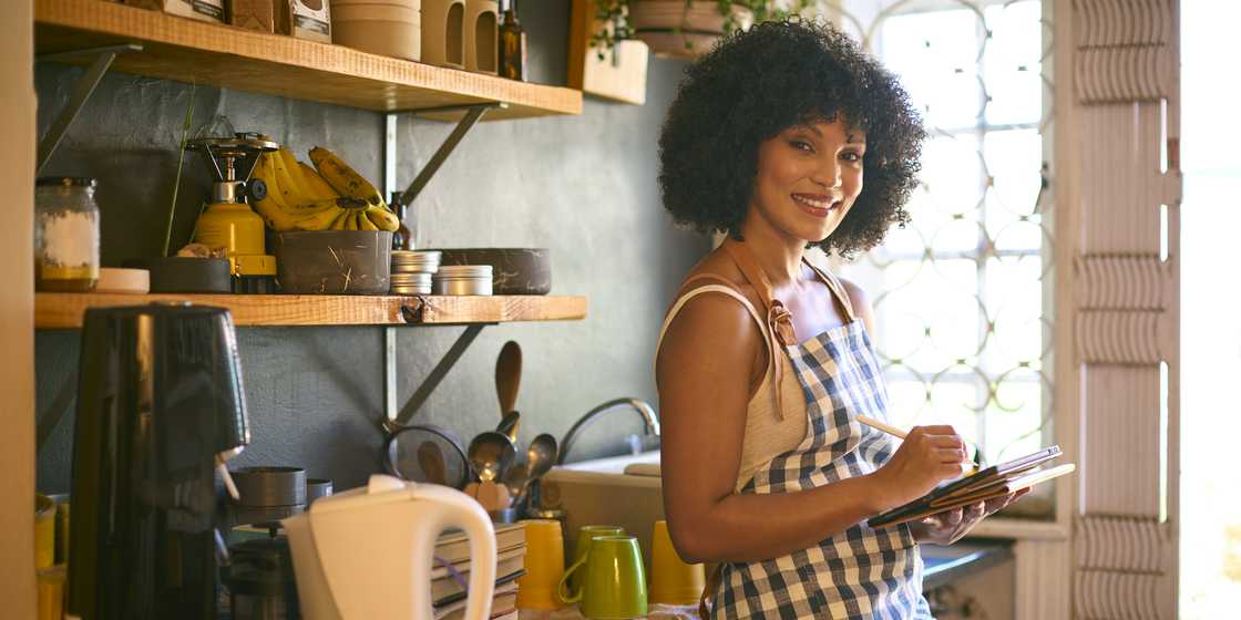 Person in a checkered apron smiles while writing in a notebook in a cozy kitchen.